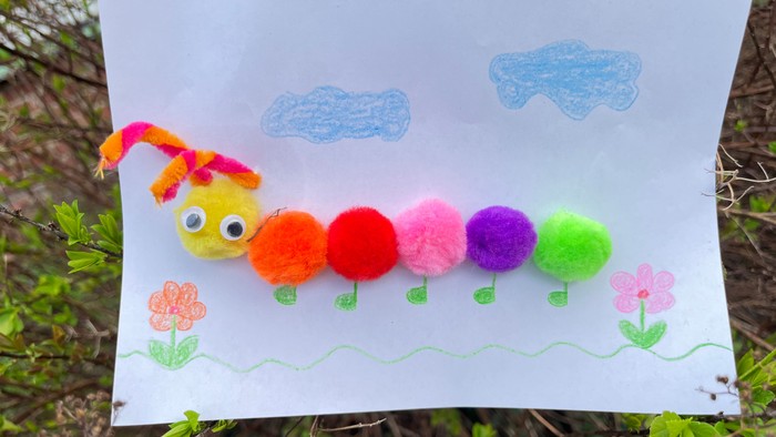 Die Abbildung zeigt eine Raupe aus bunten Pompons, die auf weißes Papier geklebt ist. Am Kopf befinden sich zwei Fühler aus bunten Pfeifenputzern und zwei Wackelaugen. Die Beine sind aufgemalt. Einfache Zeichnungen von Wolken und Blumen umrahmen die Raupe