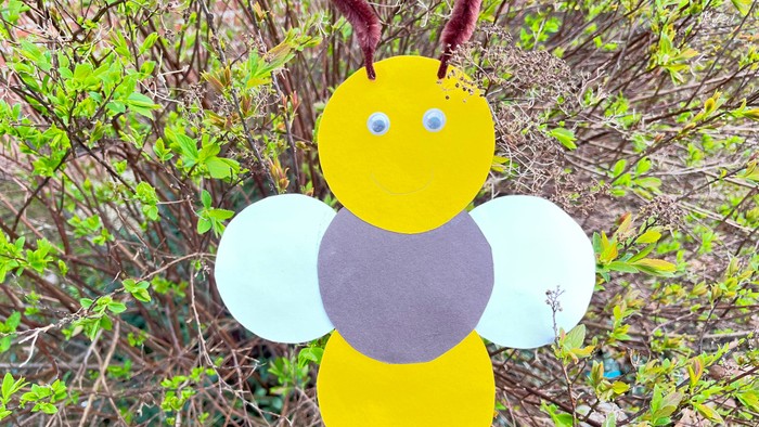 Die Abbildung zeigt eine gebastelte Biene, vor einem Hintergrund aus Zweigen und grünen Blättern, aus gelben, braunen und weißen Kreisen, die miteinander verklebt sind. Die Fühler sind aus braunen Pfeifenputzern.