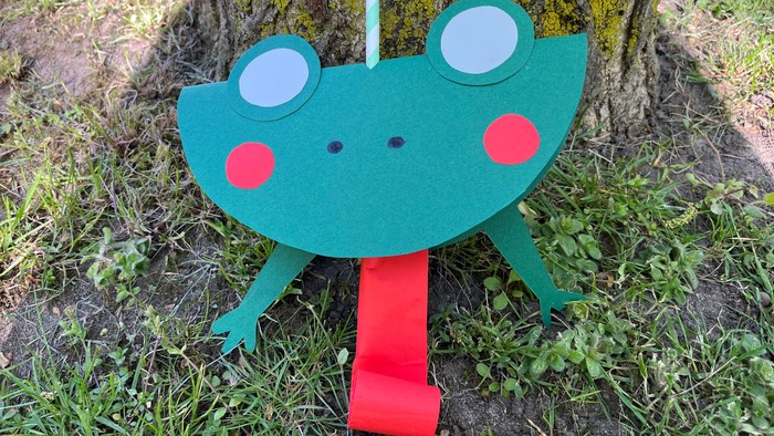 Auf der Abbildung ist ein selbst gebastelter Frosch auf einer Wiese vor einem Baum zu sehen. Der Frosch ist aus grüner Pappe geschnitten. Aus dem Mund kommt eine lange rote Zunge, die sich bei pusten in einen Strohhalm (steckt oben im Kopf) aufrollt.