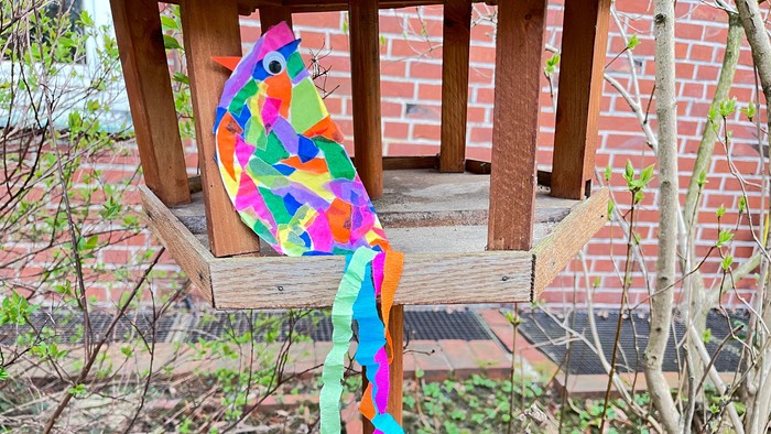 Die Abbildung zeigt einen bunten Vogel aus einem Pappteller als Körper, der mit bunten Schnipseln beklebt ist. Der Schwanz des Vogels besteht aus bunten langen Krepppapierstreifen. Der Vogel ist auf einem hölzernen Vogelhäuschen platziert.
