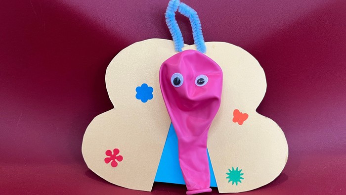 Das Bild zeigt (vor rotem Hintergrund) einen selbst gebastelten bunten Schmetterling aus Pappe. Der Körper des Schmetterlings besteht aus einem unaufgeblasenen Luftballon, der aufgeblasen werden kann, um so den Schmetterling fliegen zu lassen.