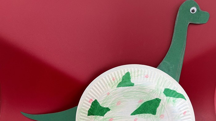 Das Bild zeigt (vor rotem Hintergrund) einen aus einem halben weißen Pappteller als Rumpf gebastelten Dinosaurier mit weiteren Körperteilen wie Beine, Schwanz und Hals mit Kopf aus grünem Tonpapier.