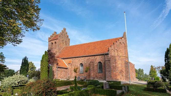 En gammel kirke med røde tag og gotiske vinduer, omgivet af græs og træer under et klart blå himmel
