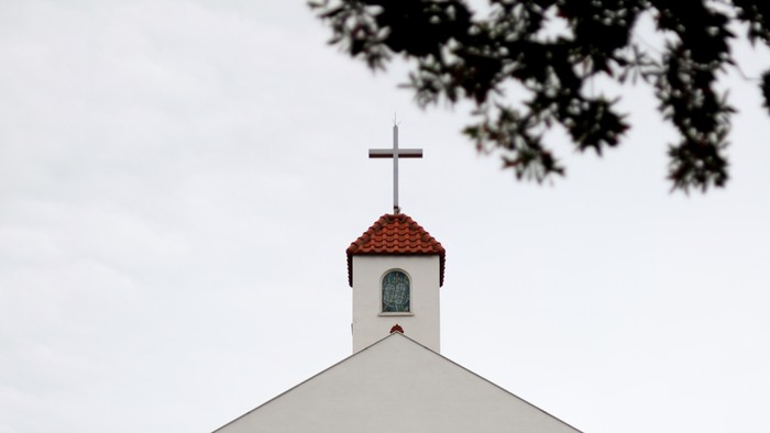 Weißer Kirchturm mit roter Kappe und Kreuz, teilweise von Bäumen verdeckt