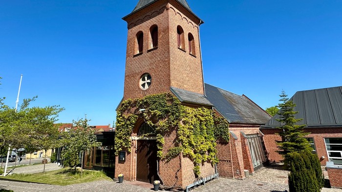 En charmerende murstenskirke med en vedbenddækket indgang og et højt klokketårn under en klar blå himmel.