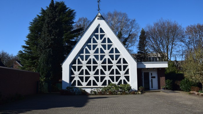 Weiße Kirche mit geometrischem Muster an der Fassade, umgeben von Bäumen und einem Kopfsteinpflasterweg.