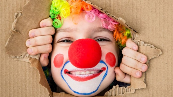 Børn med klovnsmask og regnbuehår ser ud af papkass.