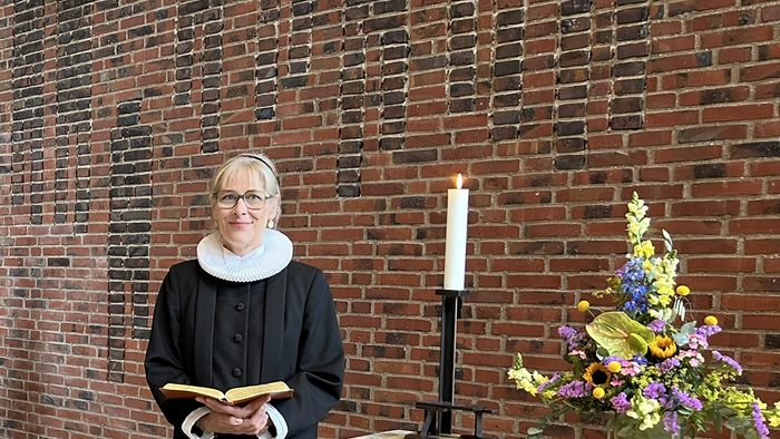 En kvinde i svart kapittel holder en bog og står foran en mur af røde mursten.