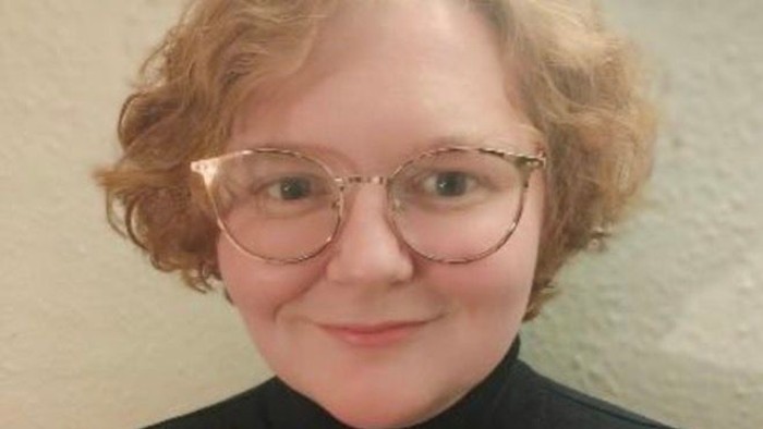 Foto af Sande Frydenlund, med kort bølget rødblond-pagefrisure, store briller med lyst hornfarvet stel, og sort trøje