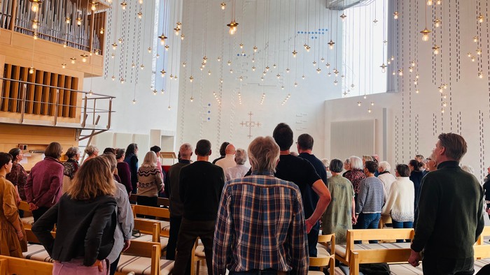 En menighed samles i en moderne kirke med hængende lysarmaturer og træbænke under en gudstjeneste.