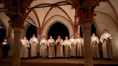 Eine Gruppe von Sängern in Roben tritt in einem schwach beleuchteten, prunkvollen Kreuzgang auf und hält Kerzen.