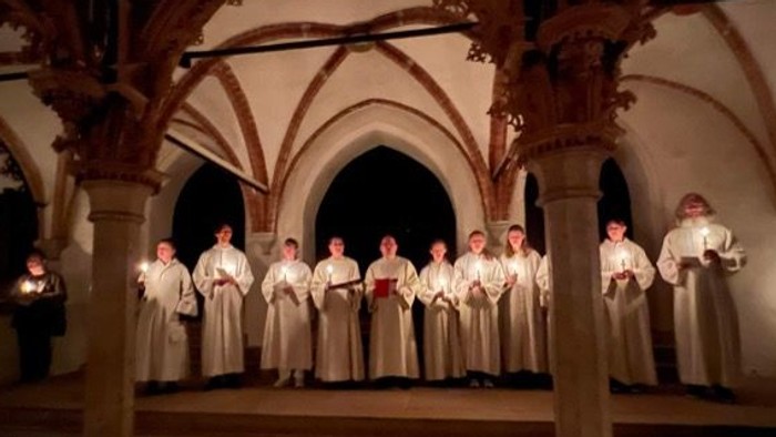 Eine Gruppe von Sängern in Roben tritt in einem schwach beleuchteten, prunkvollen Kreuzgang auf und hält Kerzen.