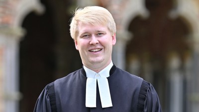 Eine lächelnde Person in akademischer Robe und Kollar posiert im Freien.