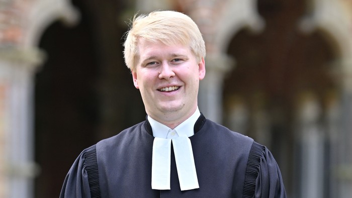 Eine lächelnde Person in akademischer Robe und Kollar posiert im Freien.