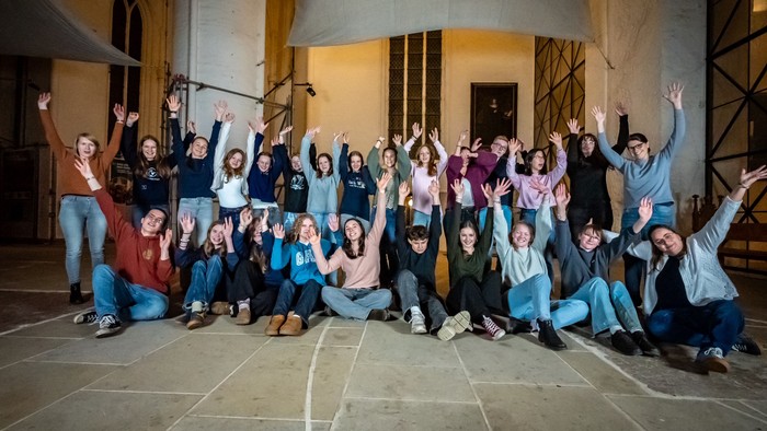 Eine fröhliche Gruppe posiert glücklich mit erhobenen Armen vor dem Eingang eines architektonischen Gebäudes.