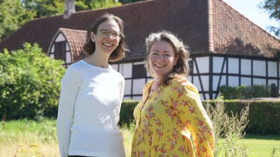 To kvinder smiler foran et hus med stråtag.