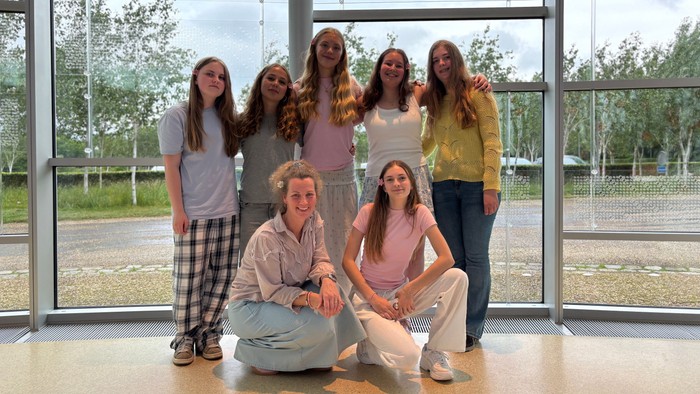 En gruppe af syv unge piger poserer sammen i et lys og moderne indgangsområde med store glassdøre og grønne omgivelser.