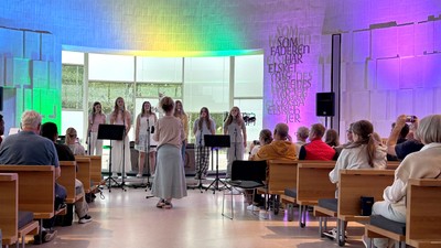 Et kor optræder på scenen i et moderne, farverigt auditorium med et opmærksomt publikum siddende foran.