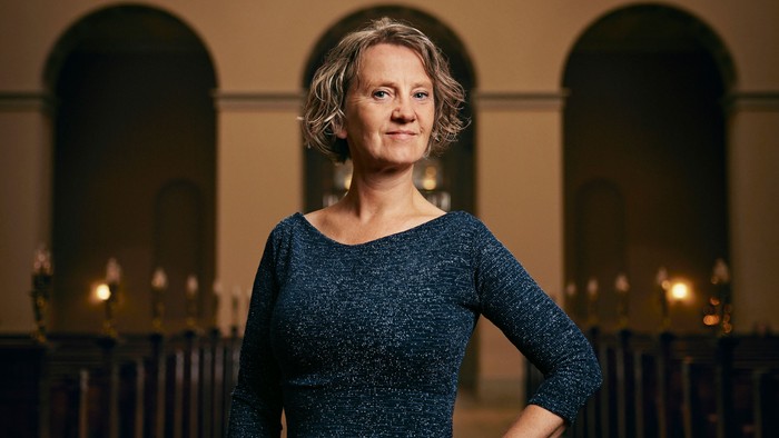 En kvinde i en mørk kjole står selvsikkert foran et prægtigt orgel i en storslået arkitektonisk omgivelse.