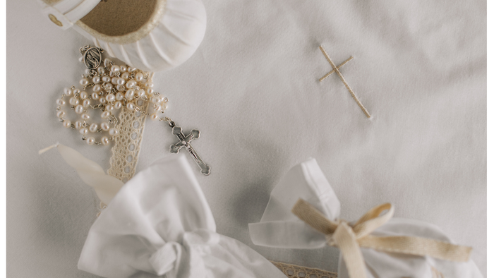 A white cloth embroidered with a cross; on top the cloth is a child's white satin shoe, the sleeve of a garment, a white tied pouch and a white rosery.