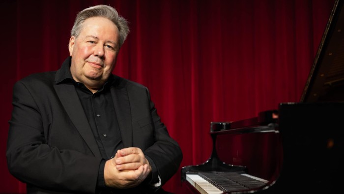 En mand i mørk suite sidder ved et piano foran rød teaterkulisse