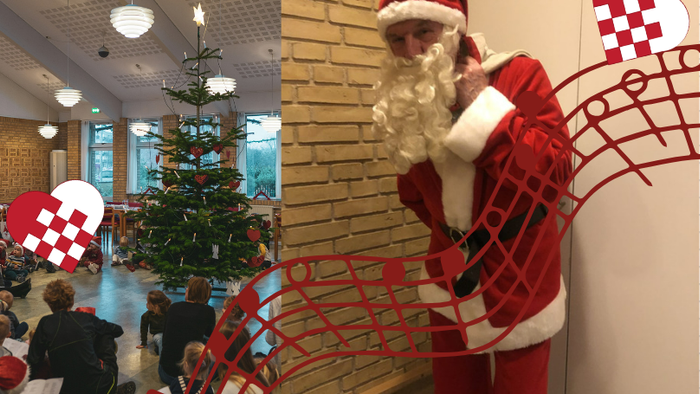 Santa Klaus står ved en mur med juledekorationer og en juletree.