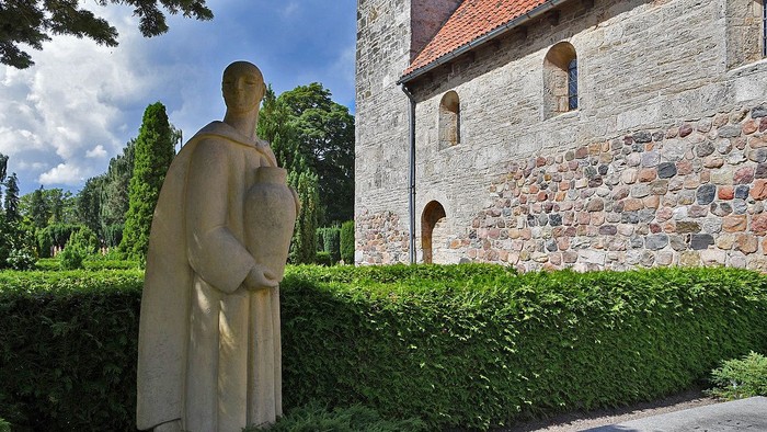 En statue af en person med skjold står foran et stenhus med rundbuefænster.