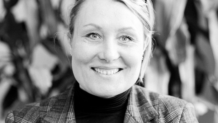 En glad kvinde med briller og en ternet blazer smiler udendørs mellem løv.