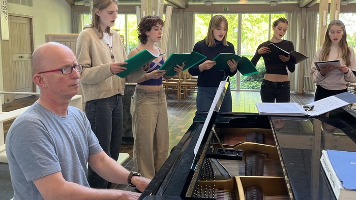 En gruppe sangere øver sammen med en pianist i et rummeligt, veloplyst musiklokale.