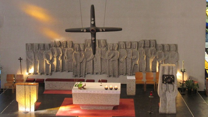 Ein großes Kreuz hängt von der Decke über einem Altar, auf dem drei Kerzen brennen. Links vom Altar befindet sich der Ambo, rechts steht der Tabernakel.
