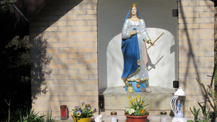 Eine bekrönte Marienstatue in einer gemauerten Nische, umgeben von Blumenstöcken und Grablaternen.