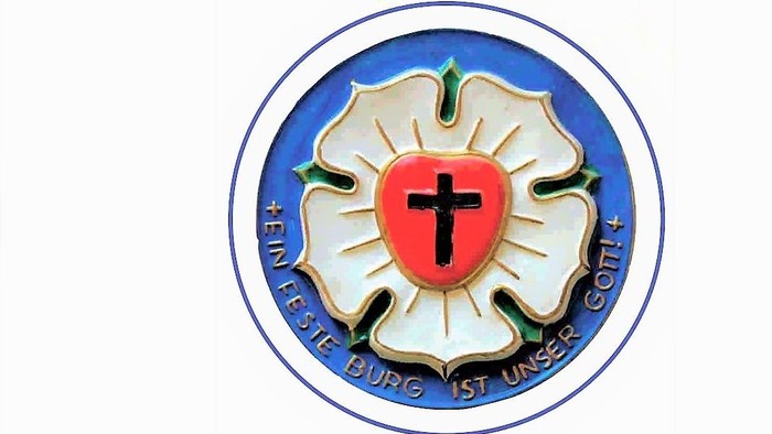 Das Bild zeigt ein Emblem mit einem roten Kreuz in einem weißen Schild, umgeben von einem blauen Ring mit deutschem Text