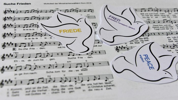 Notenblatt mit ausgeschnittenen Friedenstauben, beschriftet mit „FRIEDEN“ und „PEACE“, überlagert auf deutschen Liedtexten.