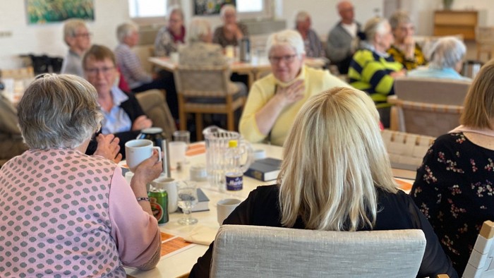 En gruppe ældre personer sidder ved bord i en common room og deltager i en social aktivitet.