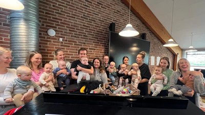 En gruppe af voksne og børn sidder sammen ved et piano i et rum med mur af røde teglsten.