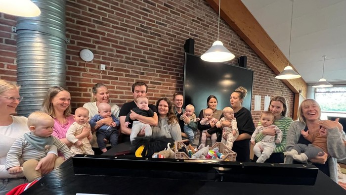 En gruppe af voksne og børn sidder sammen ved et piano i et rum med mur af røde teglsten.