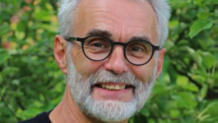 En smilende, eldre mand med briller og graat barbe står foran et grønt buskage.