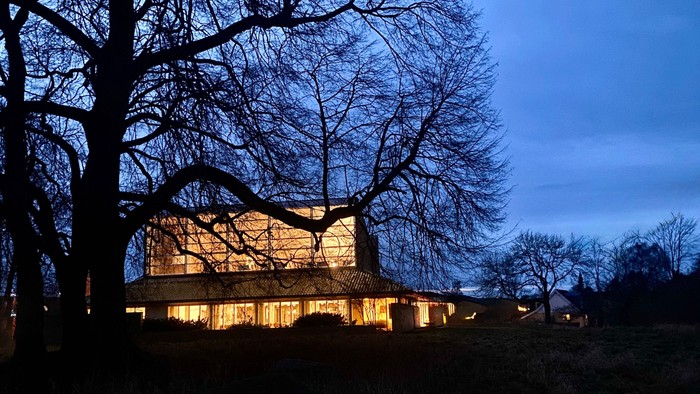 Et stort hus med mange vinduer, oplyst om natten, omgivet af bare træer.