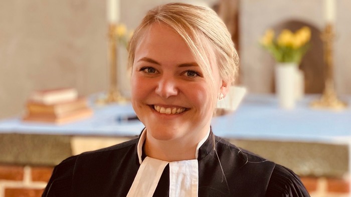 Eine Frau in schwarzer Robe mit weißem Kragen lächelt in einem Kirchenraum.