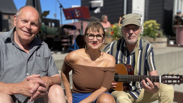 Tre personer sidder udendørs og nyder musik og samtale i solskinsvejr med en guitarist.