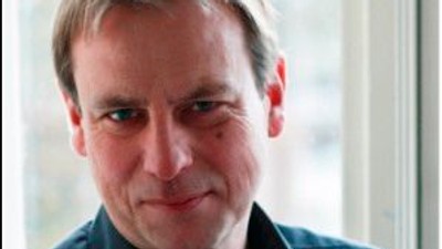 En mand med kort hår og blå skjorte står foran et vindue.