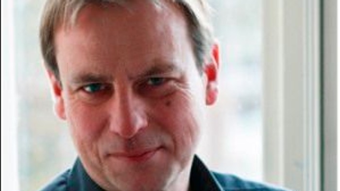 En mand med kort hår og blå skjorte står foran et vindue.