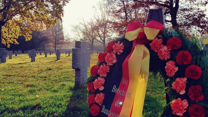Ein Blumenkranz mit roter Schleife liegt auf einem Grab in einem Friedhof.