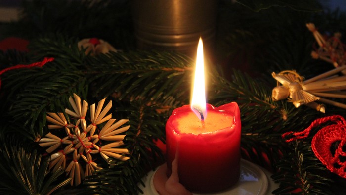 Eine brennende rote Kerze steht auf einem weißen Teller, umgeben von Grünpflanzen und Tannenzapfen.