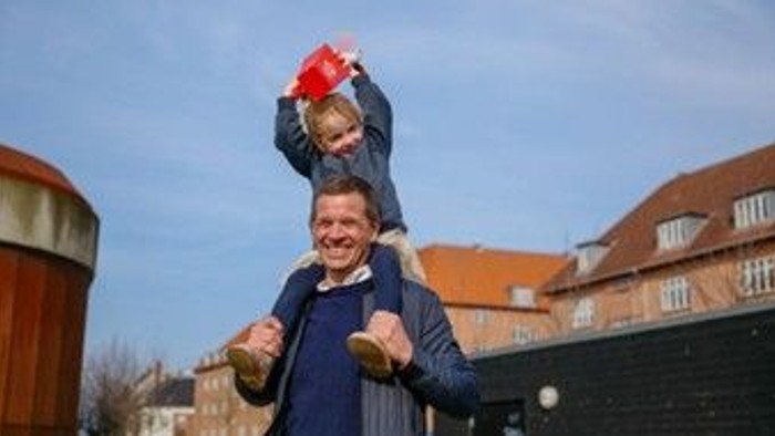 En mand løfter glædestrålende et barn højt op over hovedet, mens han holder et rødt flag.