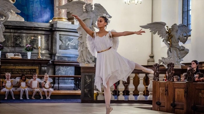 En ballerina i hvid kostume udfører en balanserende pose i en prægtig sal.