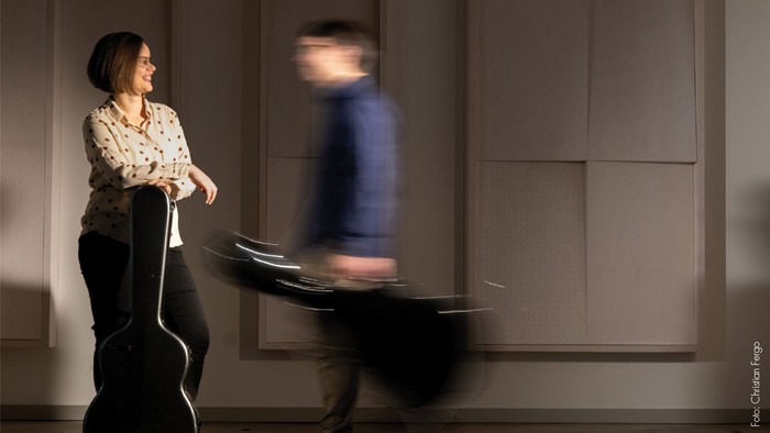 Eine Frau posiert mit einem Cello, während eine sich bewegende Person hinter ihr in einem schwach beleuchteten Raum verschwimmt.