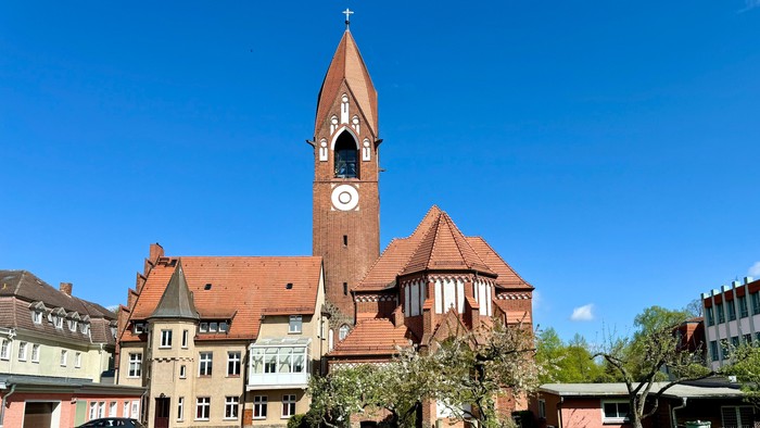 Bild zeigt eine Kirche mit hohem Turm und Uhr, umgeben von einem gepflegten Garten.