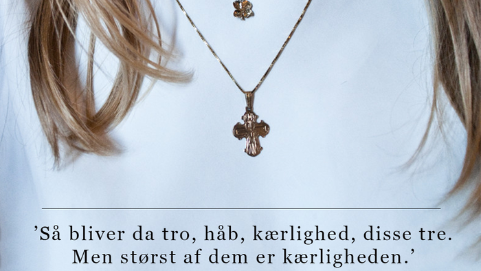 En person bærer et korshalskæde med et citat fra 1. Korintherbrev 13,13 på dansk, der fremhæver kærligheden.