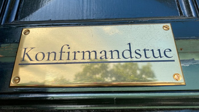 Gul bronsskilt med 'Konfirmandstue' i blå skrift.